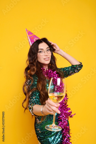 woman in a smart dress in a party cap with a pink boa around her neck and a glass of champagne smiling on a yellow background