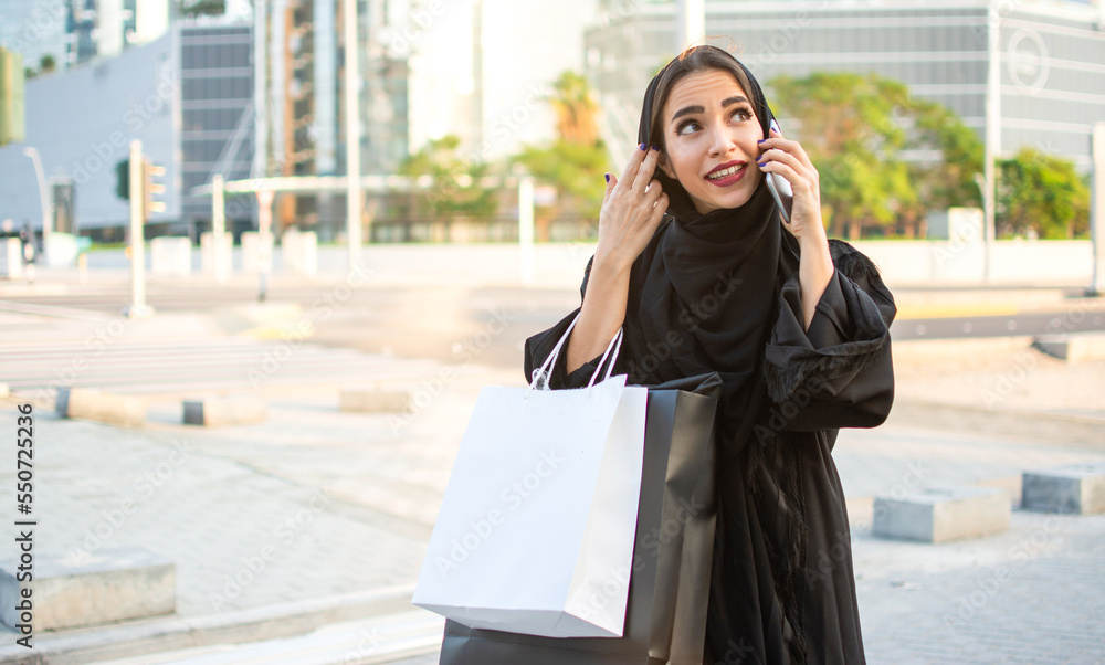 Arab woman in traditional wear holding shopping bags in hands and talking on mobile phone while standing on the street in front of the modern skyscrapers.