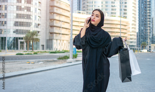 Serious Arab woman in traditional wear holding shopping bags in hands and talking on mobile phone while walking on the city street © Bojan