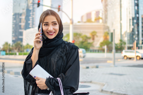 Beautiful young muslim woman in traditional clothes holding shopping bags and wallet and talking on mobile phone on the street.