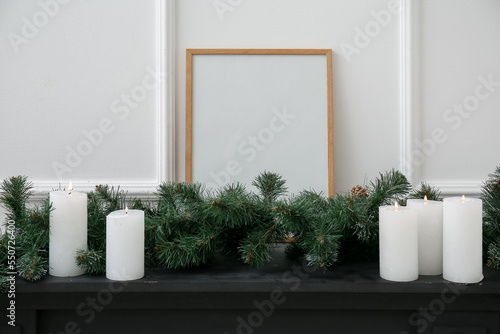 Blank frame with Christmas branches and candles on mantelpiece near light wall photo