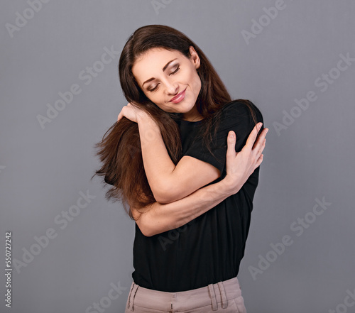 Happy calm woman hugging herself with natural emotional face in black t-shirt on grey background. Closeup