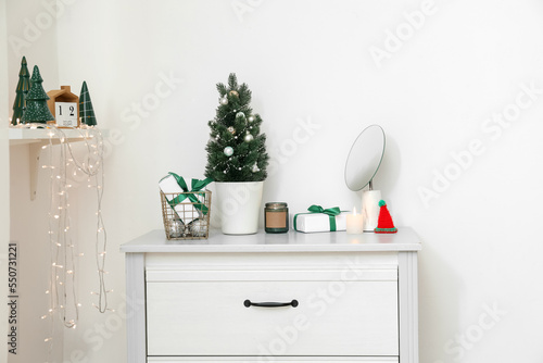 Small Christmas tree with presents, candles and mirror on chest of drawers near light wall © Pixel-Shot
