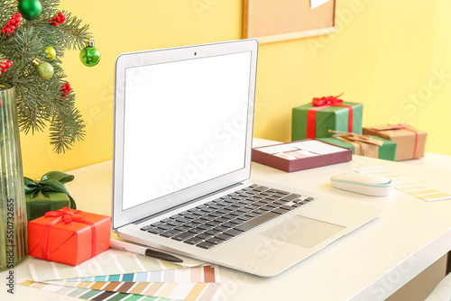 Vase with fir branches, Christmas presents and laptop on table near yellow wall
