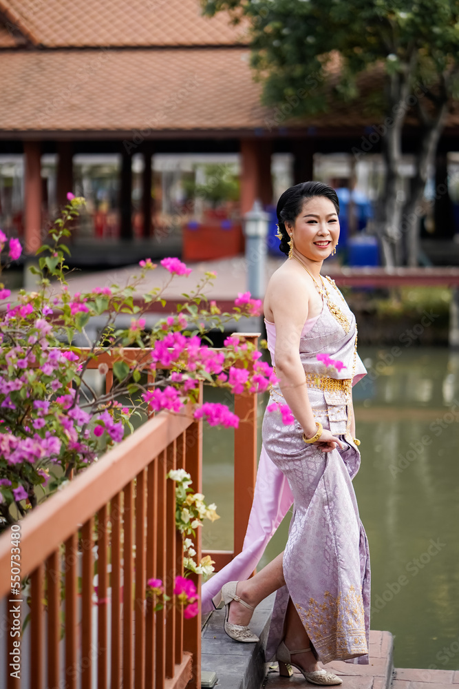 Naklejka premium A woman in a traditional Thai costumes is standing on the wooden bridge while being taken a portrait photo shooting.