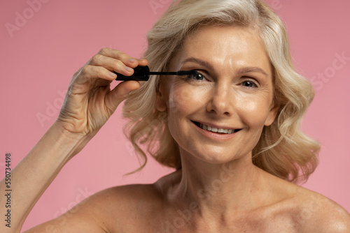 Closeup portrait of beautiful smiling mature woman applying makeup using mascara looking at mirror isolated on background. Morning routine, natural beauty concept 