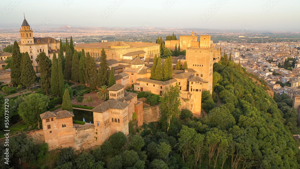 Sunrise aerial side view photo of the Alhambra fortress in Granada, Spain. The fortress is bathed in golden light.