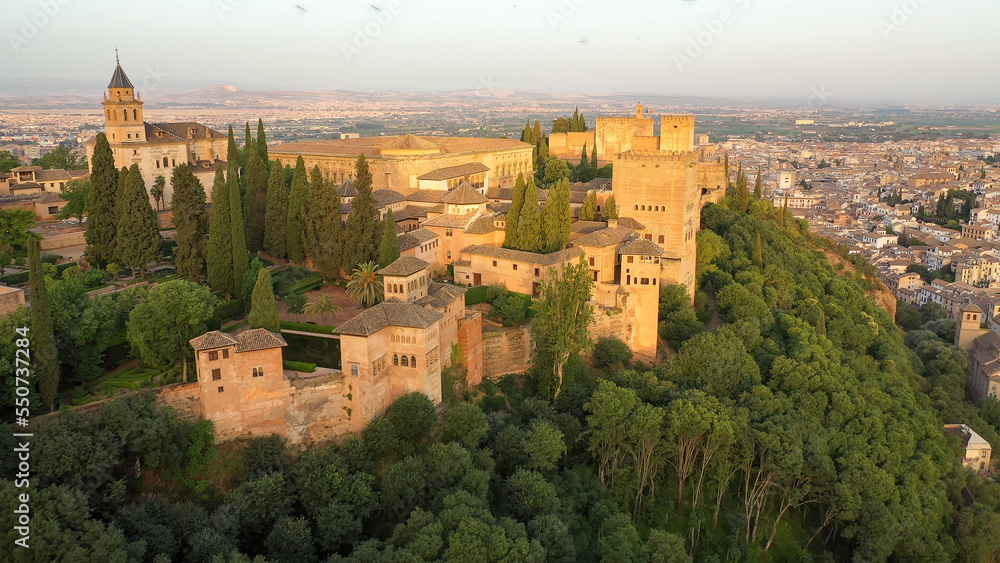 Sunrise aerial side view photo of the Alhambra fortress in Granada, Spain. The fortress is bathed in golden light.
