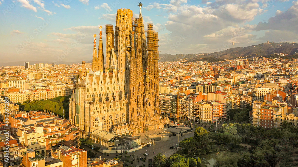 Closeup aerial view of the Sagrada Familia cathedral in Barcelona, Spain. Cathedral is bathed in golden sunrise light, blue sky, few clouds, Barcelona cityscape in background.