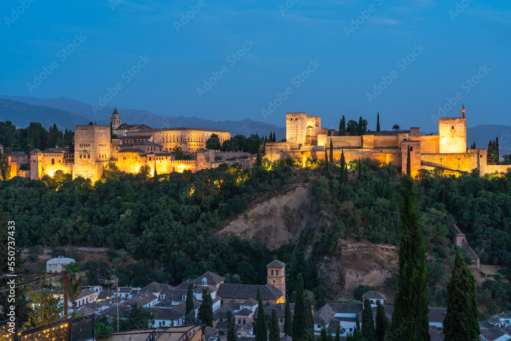 Alhambra fortress in Granada, Spain, during the blue hour. Fortress is bathed in golden light set against a cobalt blue sky.