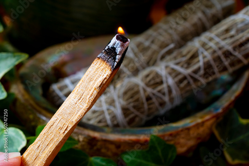 A close up image of a burning wooden smudge stick with blurred background of two white sage smudge bundles.  photo