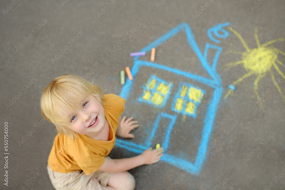 Fototapeta premium Little child boy is drawing house and sun painted with colored chalk on asphalt of sidewalk. Kids creative picture on gray background of road.