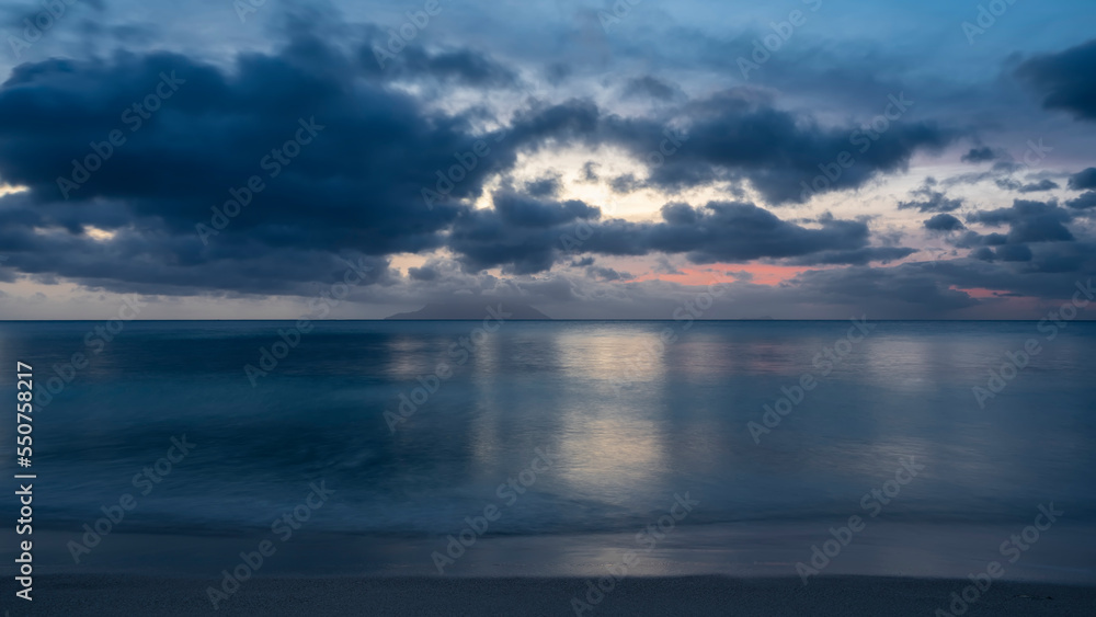 Serene evening seascape. Shades of blue. Picturesque clouds in the sky, highlighted in pink. Reflection on the water. The silhouette of the island on the horizon. Long exposure. Seychelles. Mahe Serene evening seascape. Shades of blue. Picturesque clouds in the sky, highlighted in pink. Reflection on the water. The silhouette of the island on the horizon. Long exposure. Seychelles. Mahe