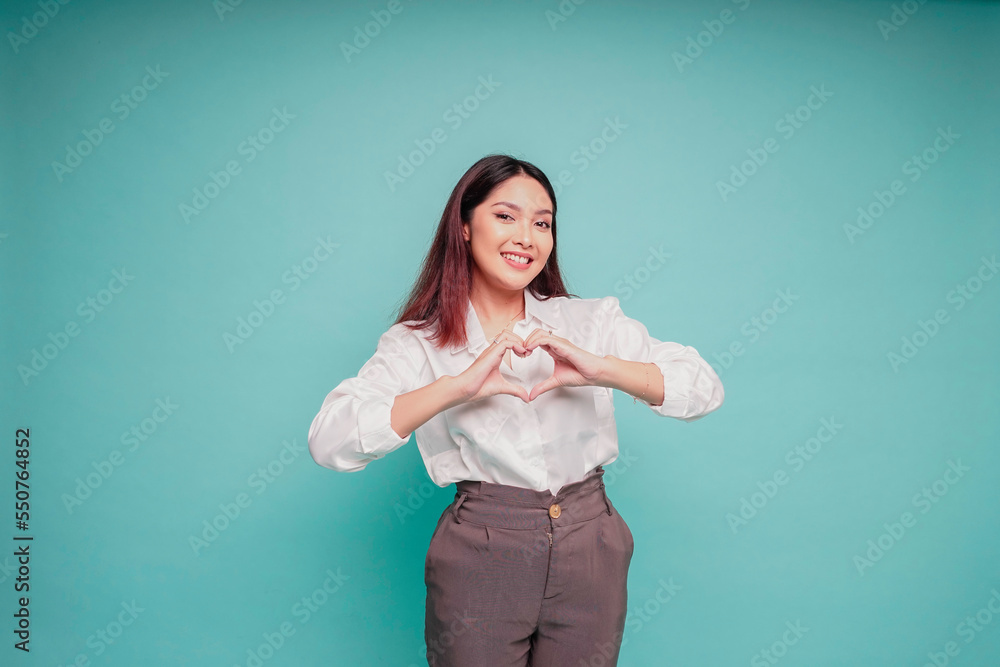 A happy young Asian woman wearing a white shirt feels romantic shapes heart gesture expresses tender feelings