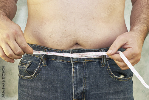 a man in jeans with a bare torso and a small belly with a measuring tape instead of a trouser belt close-up of the concept of overweight body positive