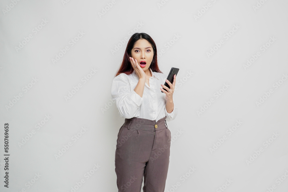 Shocked Asian woman wearing white shirt and holding her phone, isolated by white background