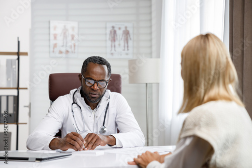 African American doctor, receives a patient, listens to complaints, makes diagnostics. Writes data. Doctor's appointment. photo