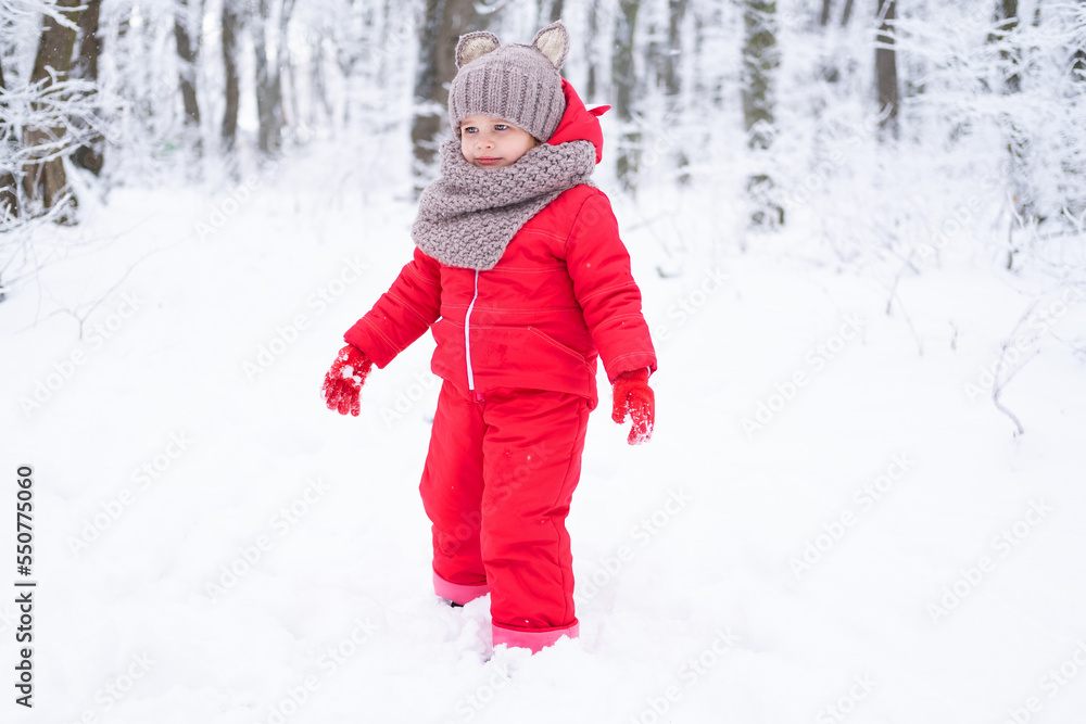 Cute little girl in pink snowsuit and knitted hat and scarf plays with snow in winter forest