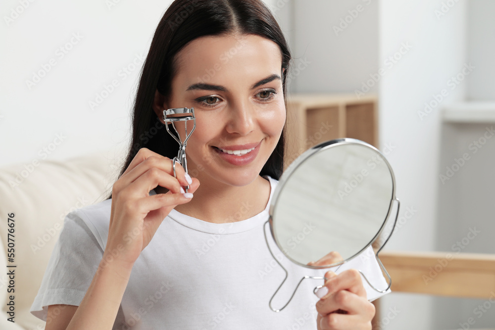 Beautiful young woman with mirror using eyelash curler indoors Beautiful young woman with mirror using eyelash curler indoors