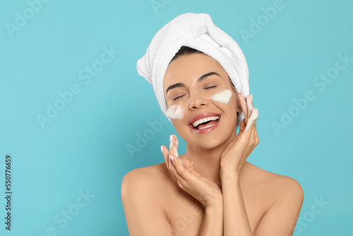 Happy young woman washing face with cosmetic product on light blue background, space for text