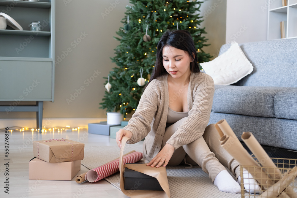 Young asian woman in beige cardigan packs gift box in wrapping paper at home, christmas time Young asian woman in beige cardigan packs gift box in wrapping paper at home, christmas time