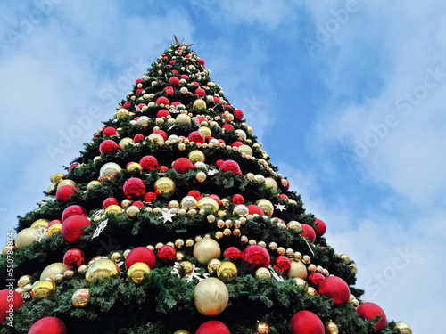 Christmas tree on the city square against the blue sky, decorated with large red and gold decorative balls and garlands. A greeting card with a place for text.