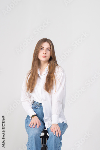 A pretty young girl in a white blouse and blue jeans sits on a stool on a white background. The model is smiling, there is a lot of copyspace around her