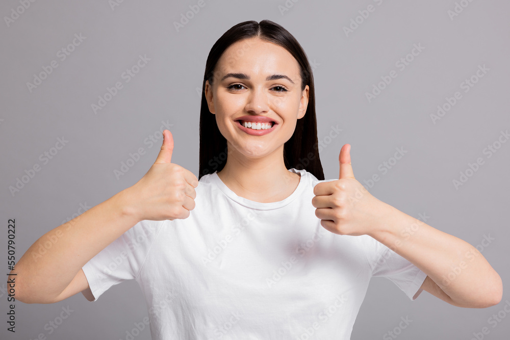Happy delighted young woman on grey background isolated showing thumb up in good mood smiling at camera wearing basic white t-shirt. Happy delighted young woman on grey background isolated showing thumb up in good mood smiling at camera wearing basic white t-shirt.
