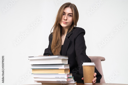 A beautiful young girl sits in front of a stack of books with coffee on a white background. The model is wearing a black jacket and trousers