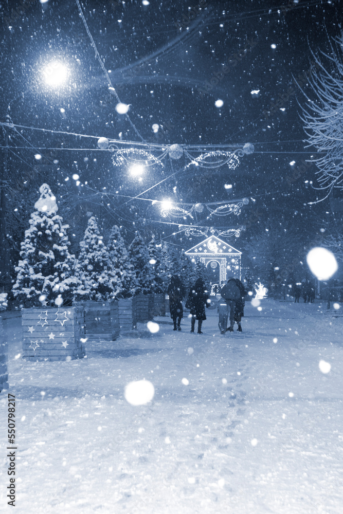 Obraz premium City street during heavy snowfall at winter night. Illumination decoration Lanterns garlands on Christmas tree. Blue color Christmas New Year Holidays celebration background. white spots of snowflakes