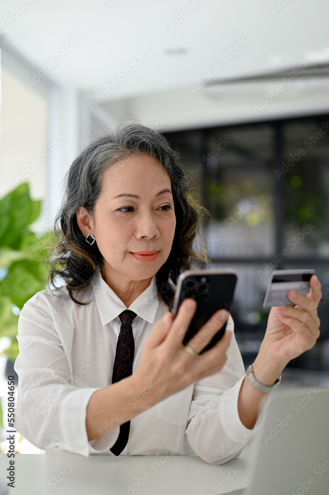 Aged Asian businesswoman holding her smartphone and a credit card, using mobile banking Aged Asian businesswoman holding her smartphone and a credit card, using mobile banking