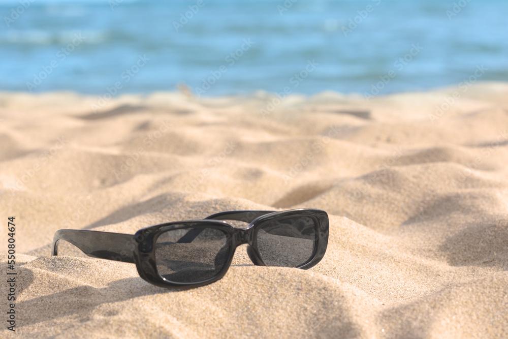 Stylish sunglasses on sandy beach near sea. Space for text
