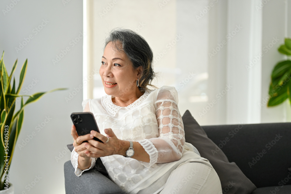 Happy Asian senior woman using her smartphone, looking at the window and daydreaming Happy Asian senior woman using her smartphone, looking at the window and daydreaming