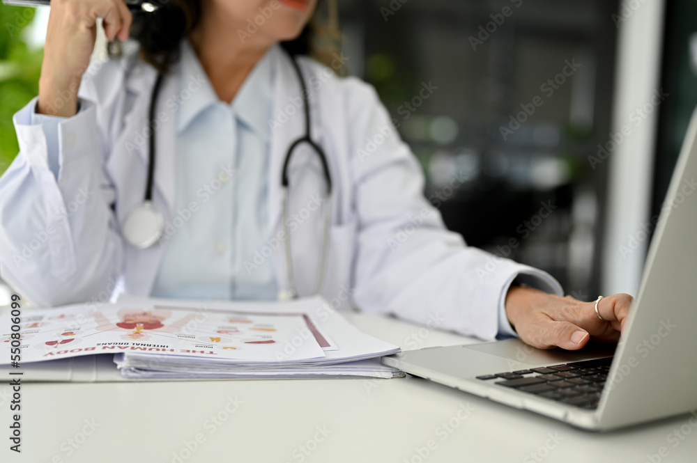 Professional Asian aged female doctor using laptop, working at her desk in the doctor's office.