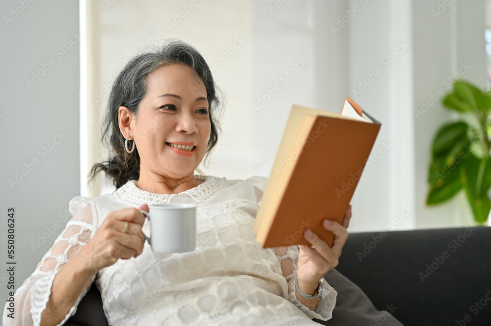 Chilling Asian aged retire woman reading a book and sipping coffee while relaxing in her living room. Chilling Asian aged retire woman reading a book and sipping coffee while relaxing in her living room.