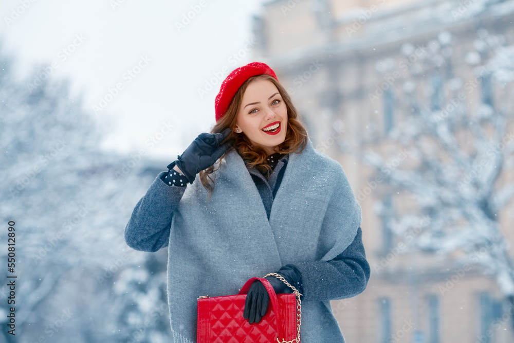 Happy smiling woman enjoying winter holidays, walking in snow covered  street of European city. Model wearing red beret, grey scarf, coat, gloves,  holding quilted bag. Copy, empty space for text Stock Photo