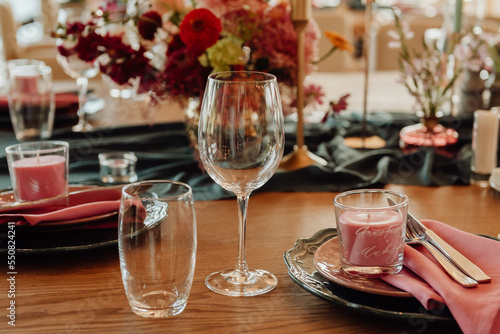 Table setting with cutlery and glass for two persons close-up. On a wooden table are a crystal goblet and a glass next to which are green plates with silver knives and forks