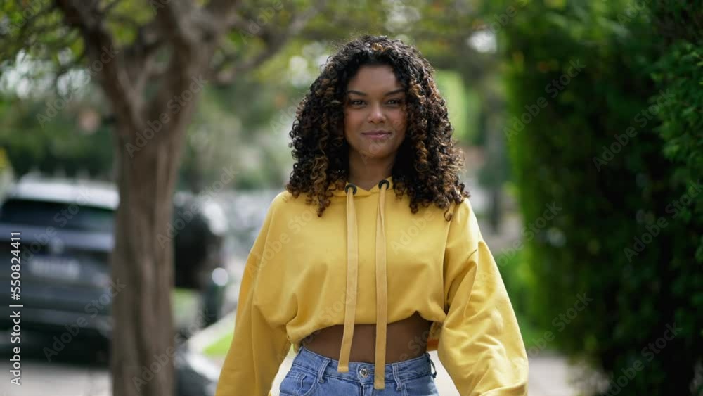A confident black girl walks forward. One happy young African American woman walking toward camera in city street sidewalk