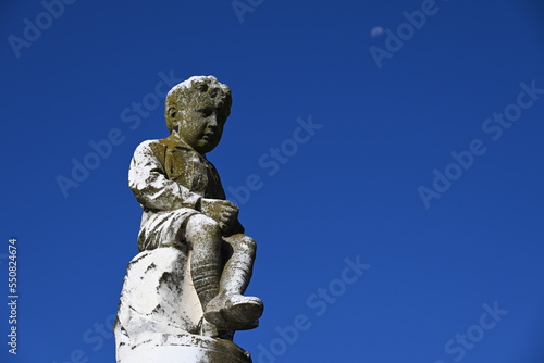 Old, weathered, and worn stone sculpture of a smartly dressed young boy seated atop a pillar in a cemetery