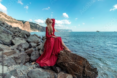 Red dress sea woman. A blonde with flowing hair in a long red dress sits on a rock near the sea. The concept of trips, a photo shoot at the sea