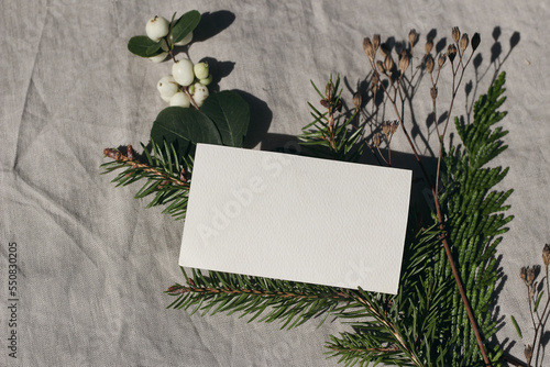 Blank textured business card mockup template. Christmas stationery, branding. Spruce, cypress tree and snowberry branch in sunlight. Beige linen tablecloth background. Selective focus. Top view. photo