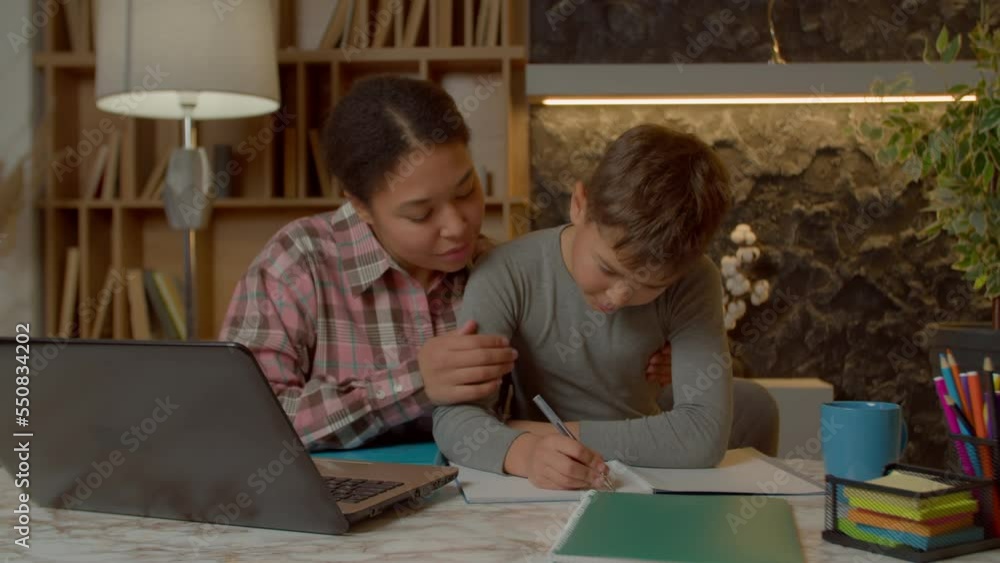 Smart adorable preadolescent multiracial boy with caring charming black mother doing homework for elementary school, writing task into textbook while studying together in domestic room. Smart adorable preadolescent multiracial boy with caring charming black mother doing homework for elementary school, writing task into textbook while studying together in domestic room.