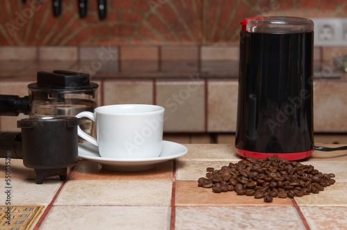 Preparing coffee: coffee beans, electric coffee grinder, coffee maker horn and white cup and saucer on a tiled kitchen table