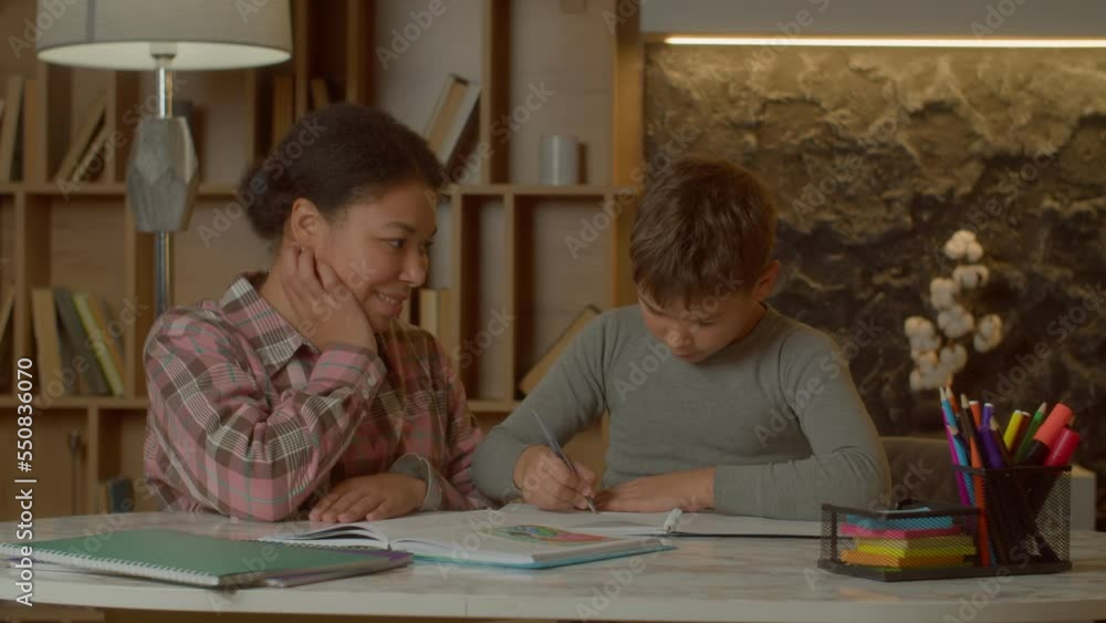 Affectionate proud pretty block mother and adorable preadolescent multicultural son chatting and bonding, expressing happiness and love while child studying homework in domestic room. Affectionate proud pretty block mother and adorable preadolescent multicultural son chatting and bonding, expressing happiness and love while child studying homework in domestic room.