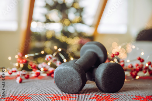 Healthy lifestyle during Christmas feasting period concept. Black dumbbells with Christmas ornaments blurred Christmas tree with Christmas lights on background, copy space.