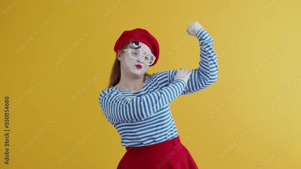 A mime girl in a striped and bright suit smiles and jokingly points to her bicep, toched it. Pantomime plays out a performance on a yellow background A mime girl in a striped and bright suit smiles and jokingly points to her bicep, toched it. Pantomime plays out a performance on a yellow background