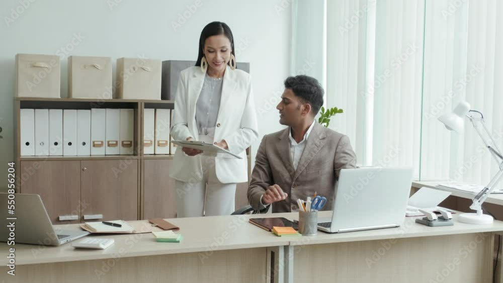 Asian businesswoman walking to male Indian colleague in office and discussing document with him while cooperating during workday Asian businesswoman walking to male Indian colleague in office and discussing document with him while cooperating during workday