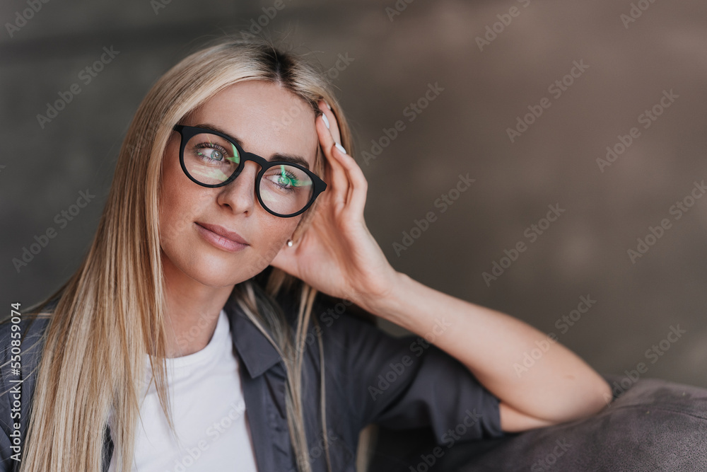Close up portrait of gorgeous blonde Swedish woman in glasses, grey shirt with long loose hair leaning on hand looks atside toothy smiles, satisfied by career of psychologist. Model posing indoors. Close up portrait of gorgeous blonde Swedish woman in glasses, grey shirt with long loose hair leaning on hand looks atside toothy smiles, satisfied by career of psychologist. Model posing indoors.