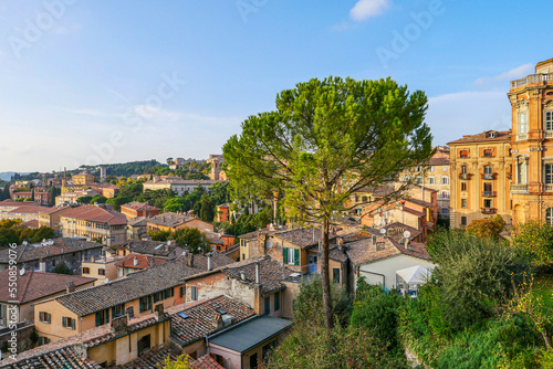 A city view from above, the mountains and the roofs of the houses can be seen. Also there is sunset, and the sun shines on some of the houses, the treetops can be seen below.