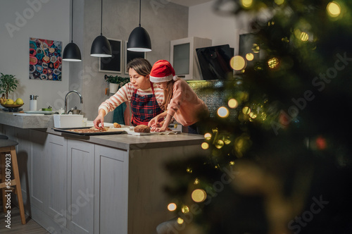 Cute little girl in red Santa hat with mother making homemade Christmas gingerbread cookies using cookie cutters together in home kitchen. Happy family holidays preparation and childhood concept.
